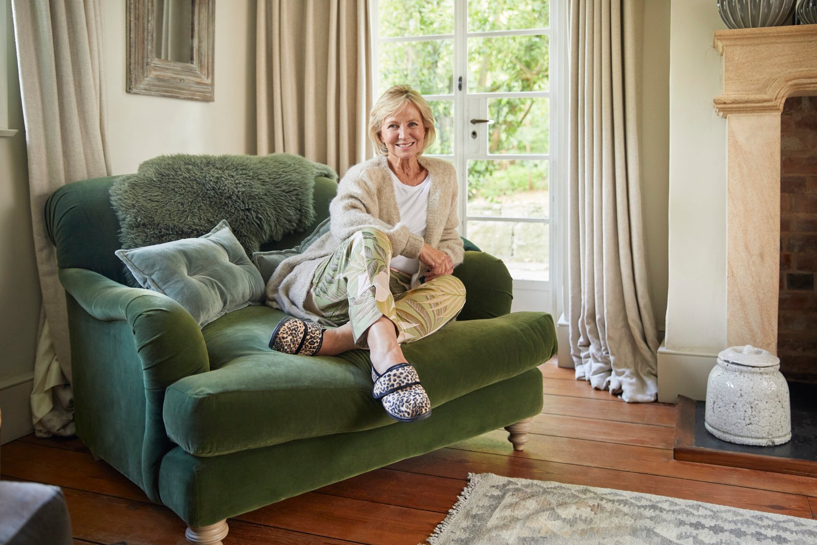 Woman sitting on a green armchair in a living room, wearing leopard print Cosyfeet Holly plush slippers.