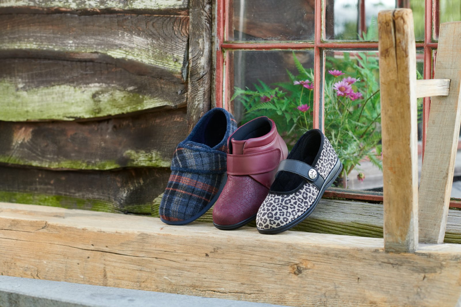 Three pairs of Cosyfeet autumn-winter footwear, including a navy plaid slipper, a claret red ankle boot, and a black and white leopard print shoe, displayed on a wooden windowsill.