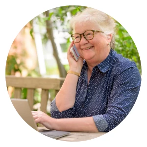 A smiling elderly woman on the phone, likely receiving customer support, with a laptop open on a table outdoors.