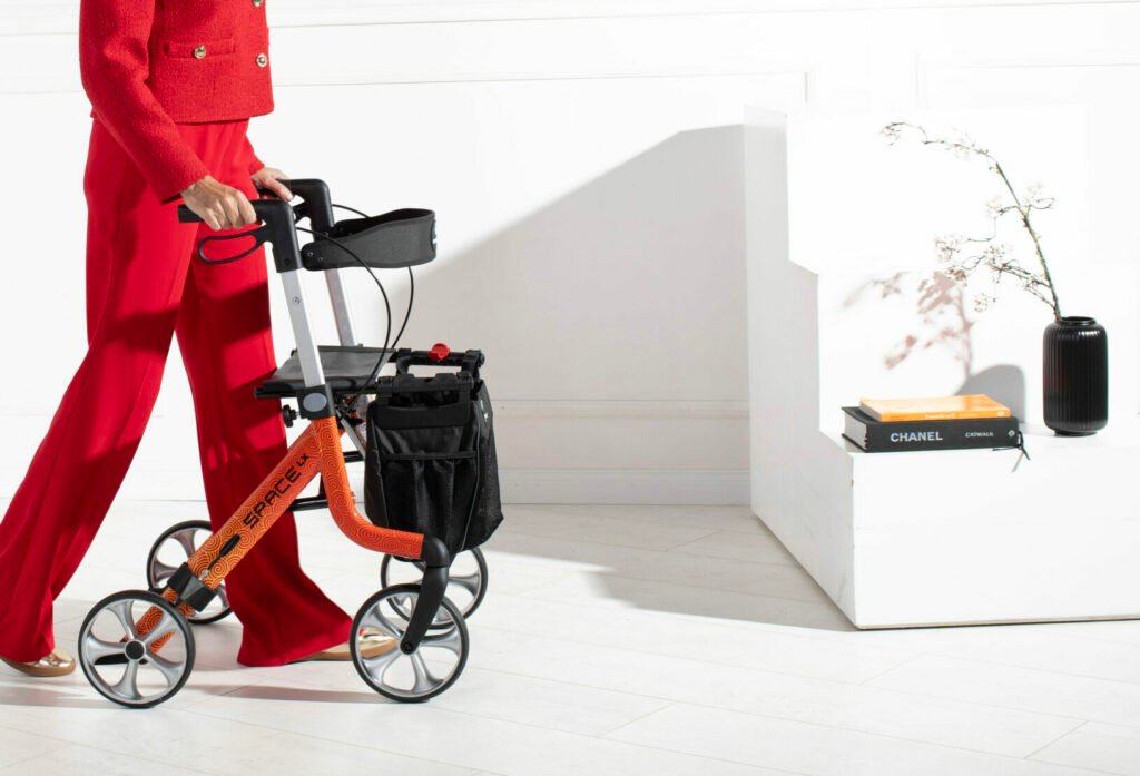 A person in red clothing using an orange Rehasense SPACE LX ART Line Rollator in Cloud design, with a stylish plant and books on a white shelf in the background.