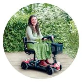 A woman smiling while riding a compact red mobility scooter outdoors in a green, leafy environment.