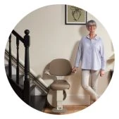 An elderly woman ascending a wooden staircase on a beige curved stairlift in a modern home.