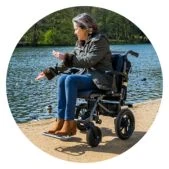 A person seated in a modern black power wheelchair next to a body of water, indicating outdoor mobility.