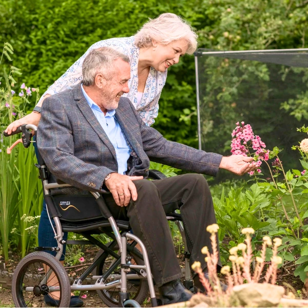 An elderly man seated in a manual wheelchair, looking at a flower, while an elderly woman gently pushes the wheelchair from behind, set in a lush garden.