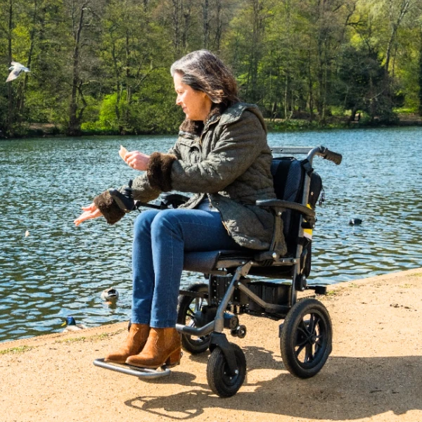 A woman in a black powered wheelchair is looking over a lake, with her arm extended as if feeding ducks.