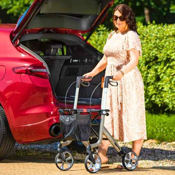 A woman in a light-coloured dress is seen placing a black Rehasense Space LX rollator into the boot of a red car, highlighting its portability.