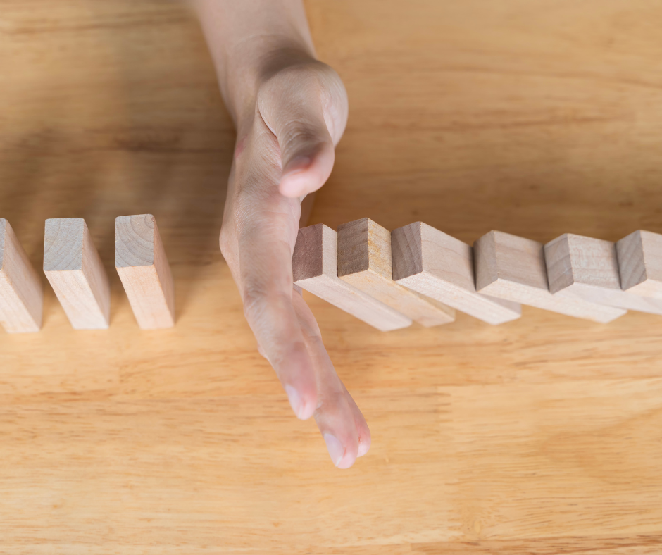 Dominos falling over on a wooden surface with a human hand preventing them from continuing to fall.