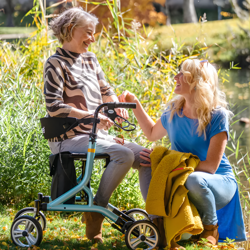 Woman seated on a light blue four-wheeled rollator, smiling at a companion kneeling beside her, enjoying a sunny day outdoors near water and reeds.