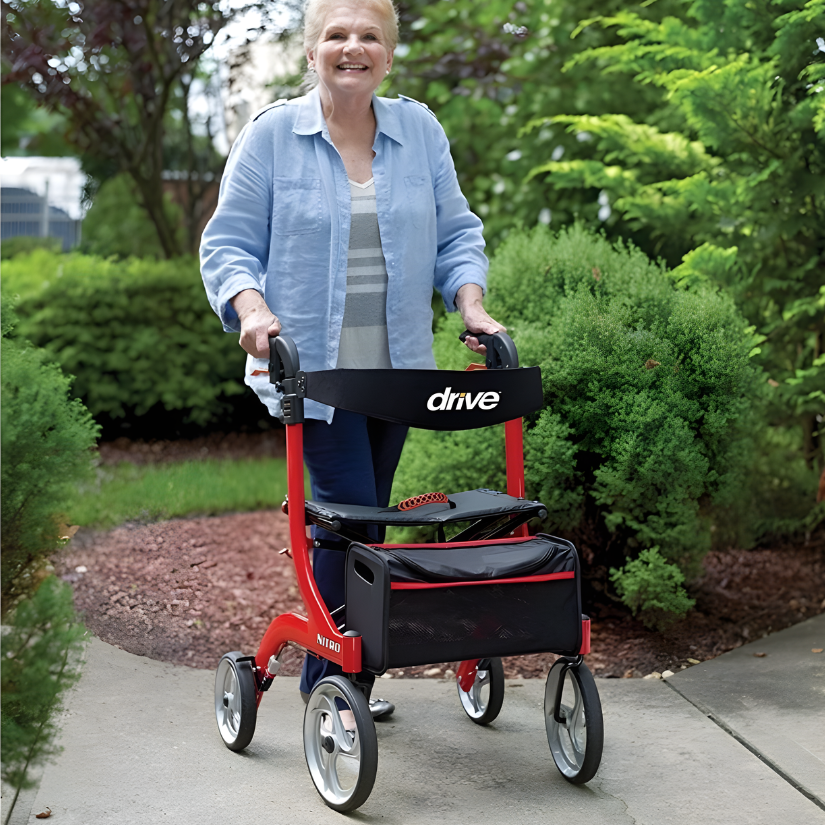 Smiling woman using a red Drive Nitro rollator with a black seat and storage bag, walking outdoors on a paved path.