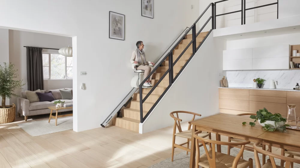 Elderly woman sitting on the Access Homeglide Stairlift, ascending a modern wooden staircase in a bright, open-plan living and kitchen area.