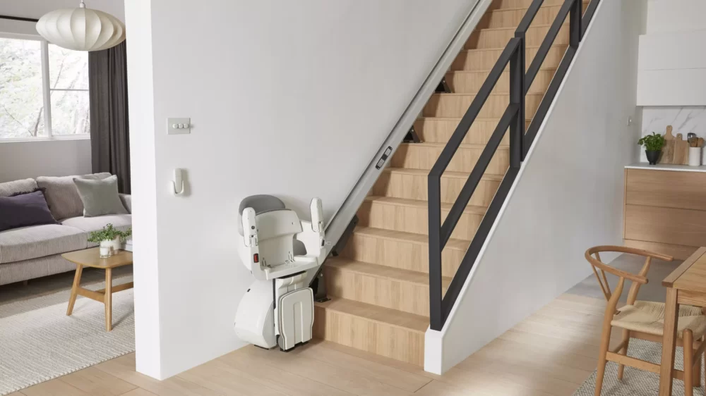 White and grey Access Homeglide Stairlift folded compactly at the base of a modern, open-riser staircase with black railings, maximizing space in the living area.