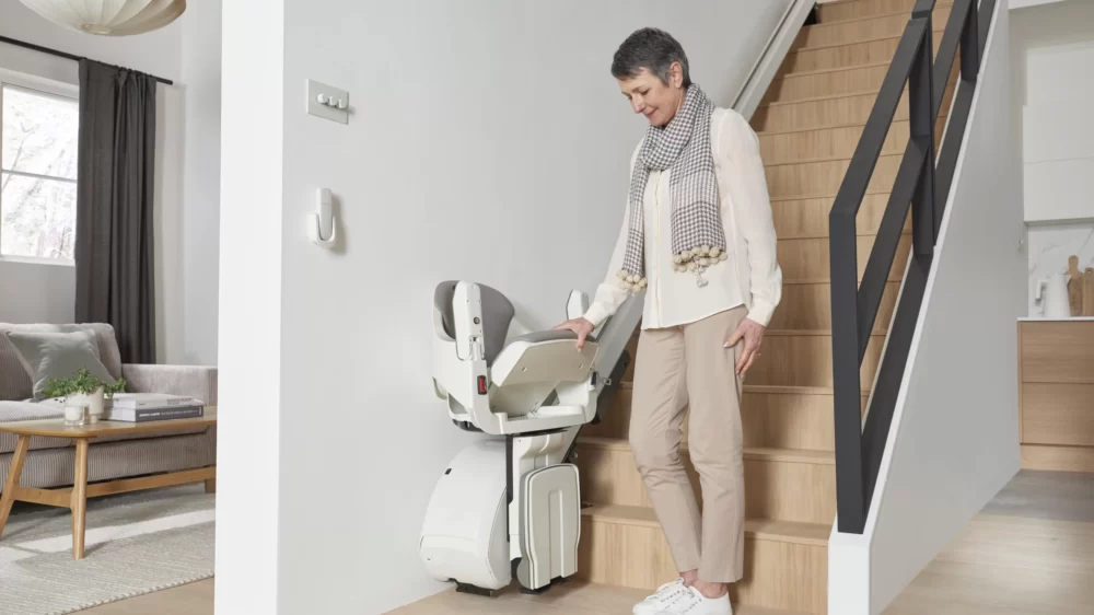 Woman standing next to the Access Homeglide Stairlift, which is folded compactly at the base of the modern staircase to maximize hallway space.