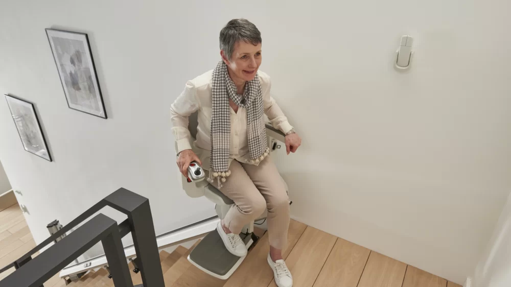 Elderly woman sitting on the Access Homeglide Stairlift, which has swiveled at the top of the stairs, demonstrating the powered swivel function for a safe dismount onto the landing.
