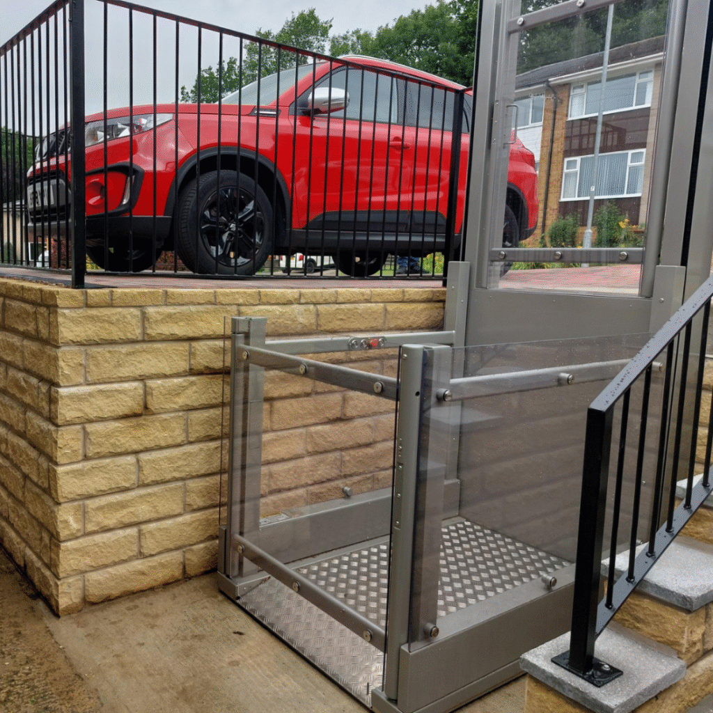 Installed Pollock Steplift Outdoor vertical platform lift on a paved residential entry, with the silver lift unit connecting the ground level to the elevated patio and parking area.