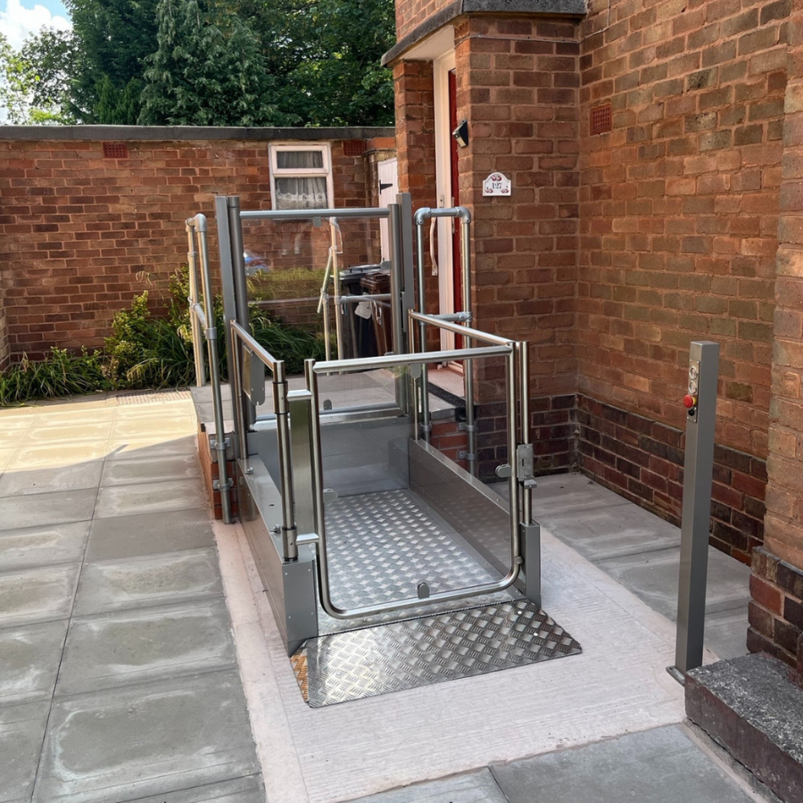 Silver Pollock Steplift Outdoor vertical platform lift installed on a paved patio next to a brick house entrance.