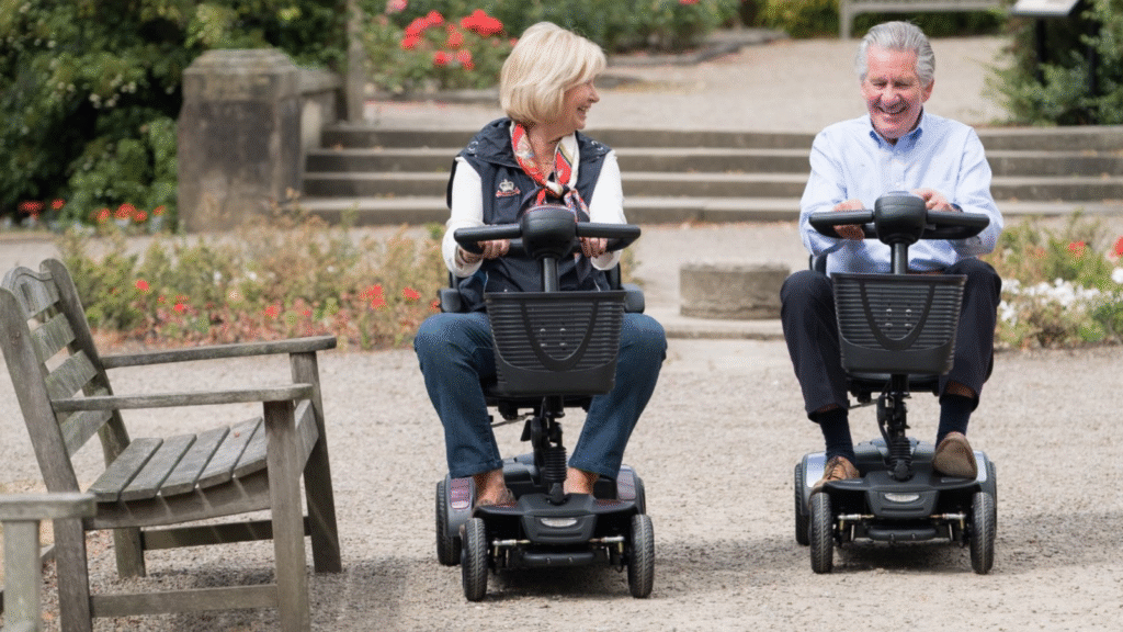 Senior man and woman smiling while riding side-by-side on two identical black pavement mobility scooters on a gravel path in a sunny park.