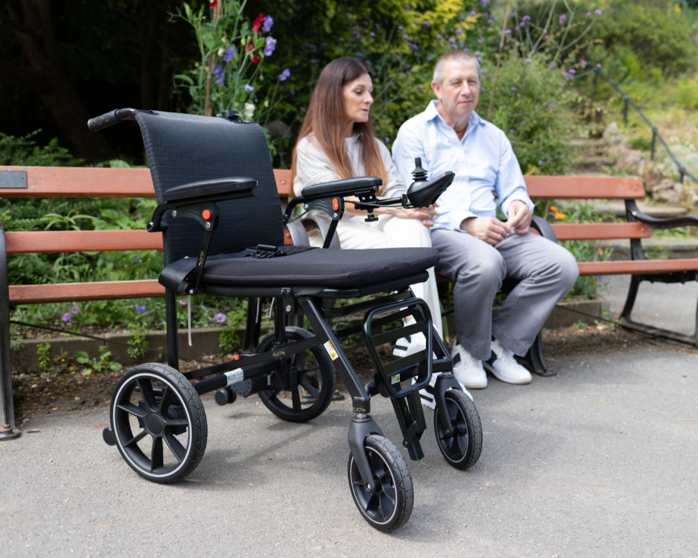 The black eFoldi Navigator foldable power wheelchair with joystick control, positioned on a paved path in a park setting near a bench.