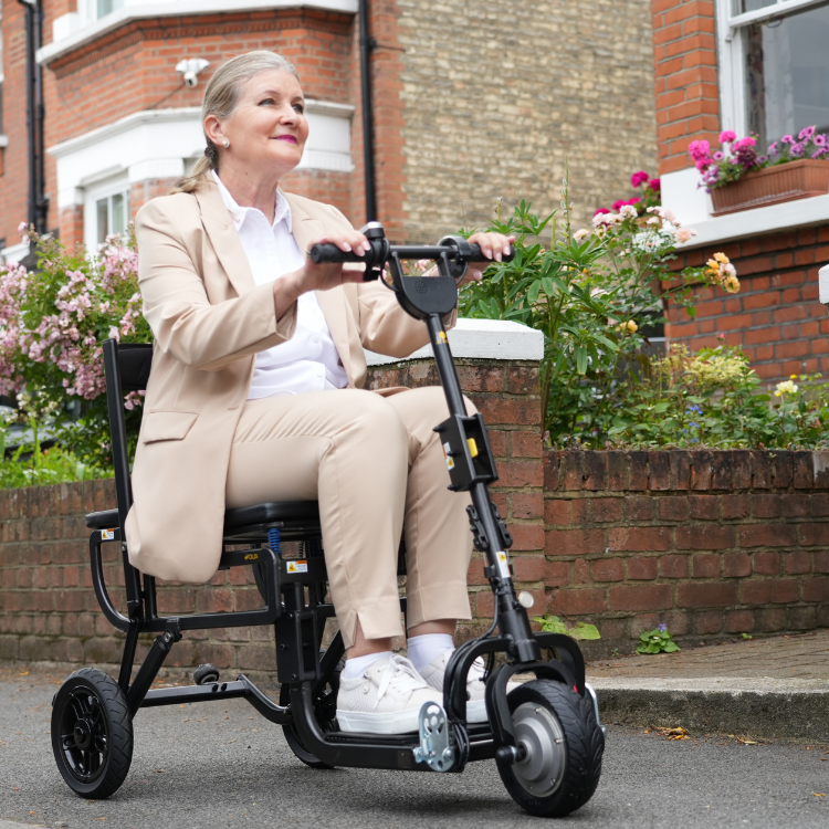 Woman riding the compact, black eFoldi three-wheeled mobility scooter outdoors on a residential street, highlighting its portability and independent use.